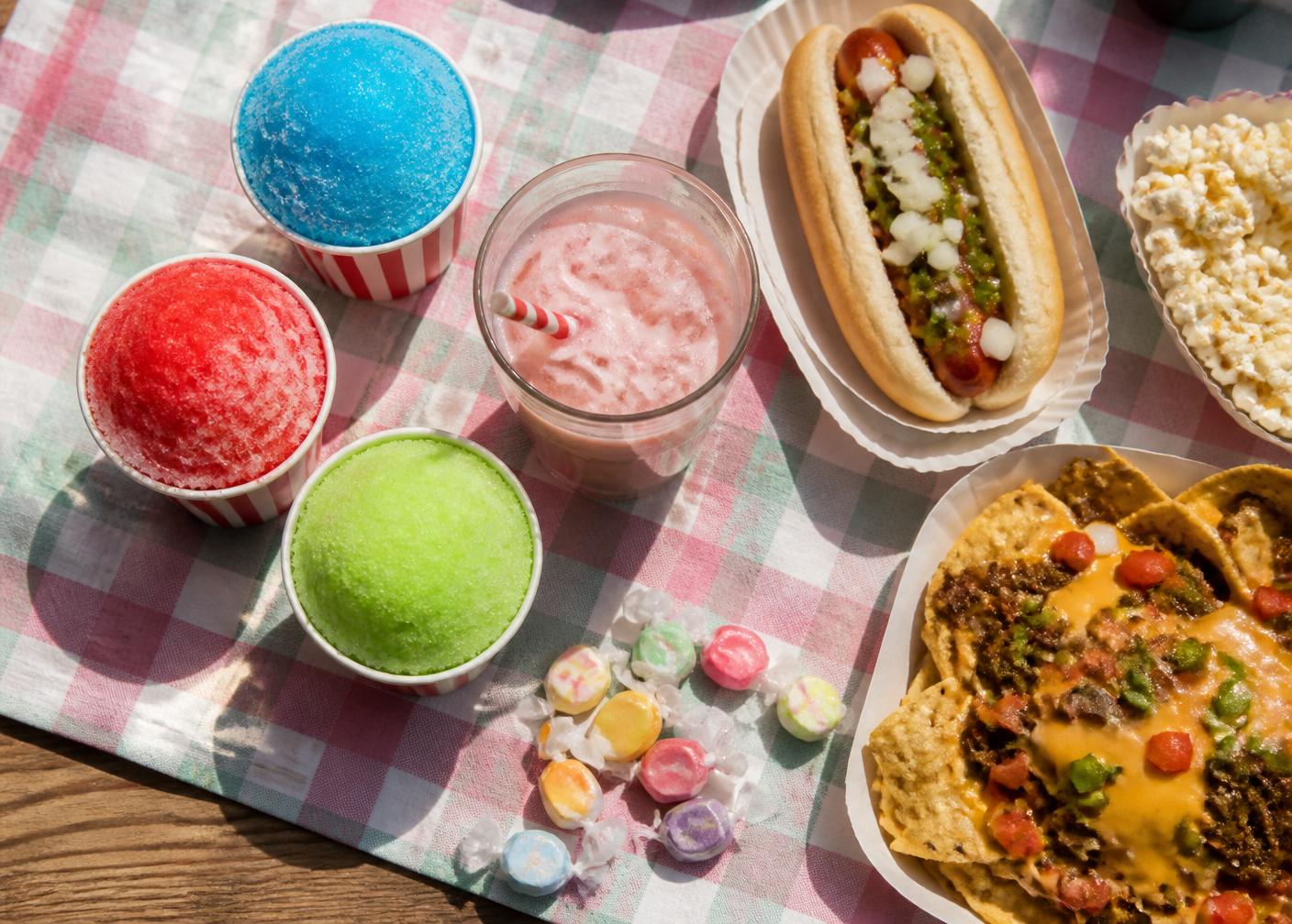 Sunny snack spread with colorful snowballs, dirty soda, hot dog, nachos, candy, and popcorn